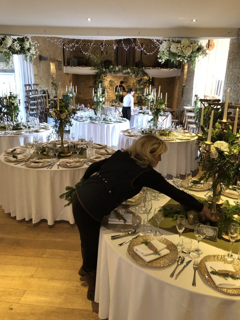 A member of our team finishing setting up tables at the great tythe barn in Tetbury with green hessian squares under a gold candelabra covered in a wreath of fake white roses and fresh season greenery surrounded but gold charger plates and a full place setting set up for hire