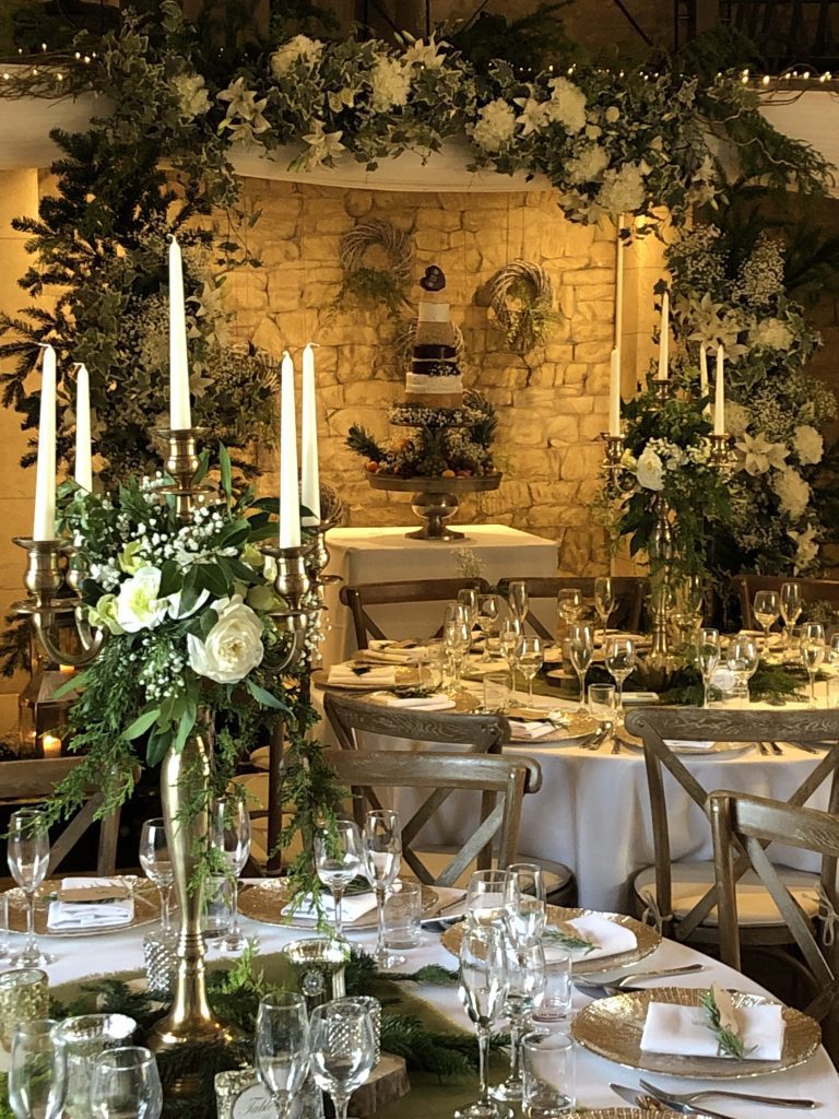 Gold candelabra, gold charger plates, and wooden slice with hessian table cloth and tealight holders in front of a white and green arch full of ivy and winter Christmas tree branches and a stacked cheese cake with rounds of Godminster cheeses including cheddar, stilton and brie for a woodland winter wedding available to hire