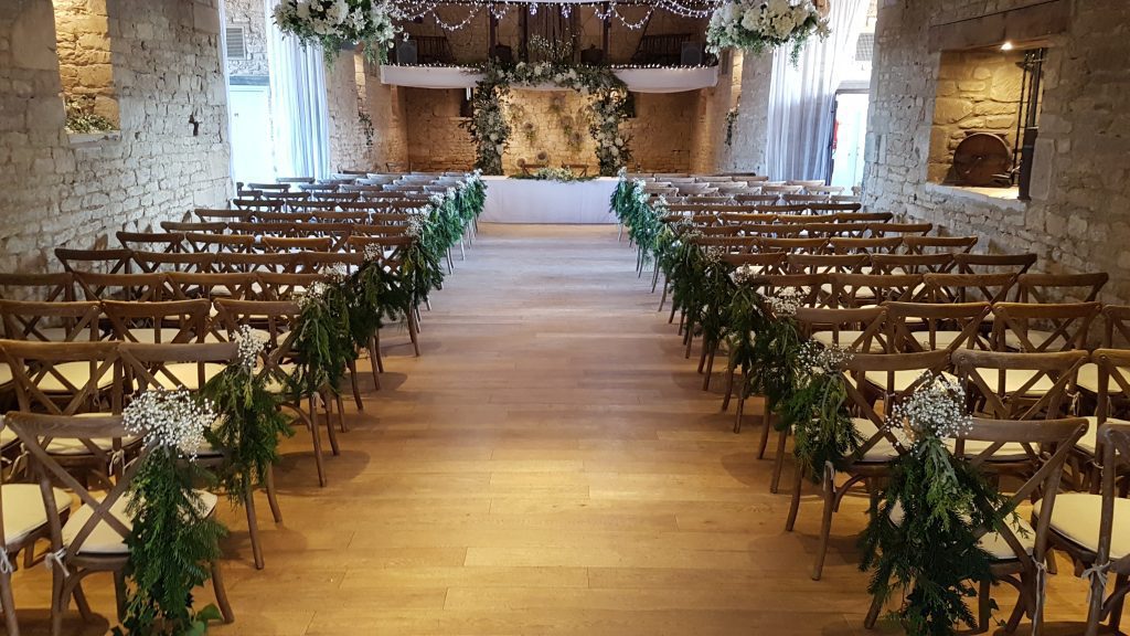 A ceremony set up at the Great Tythe barn in Tetbury with each of the aisle chair having a fern bunch and a posy of gypsophila coming out the top looking down towards a faux floral arch of winter greens and white flowers and two hanging wreaths to match along with a garland across the table all available to hire