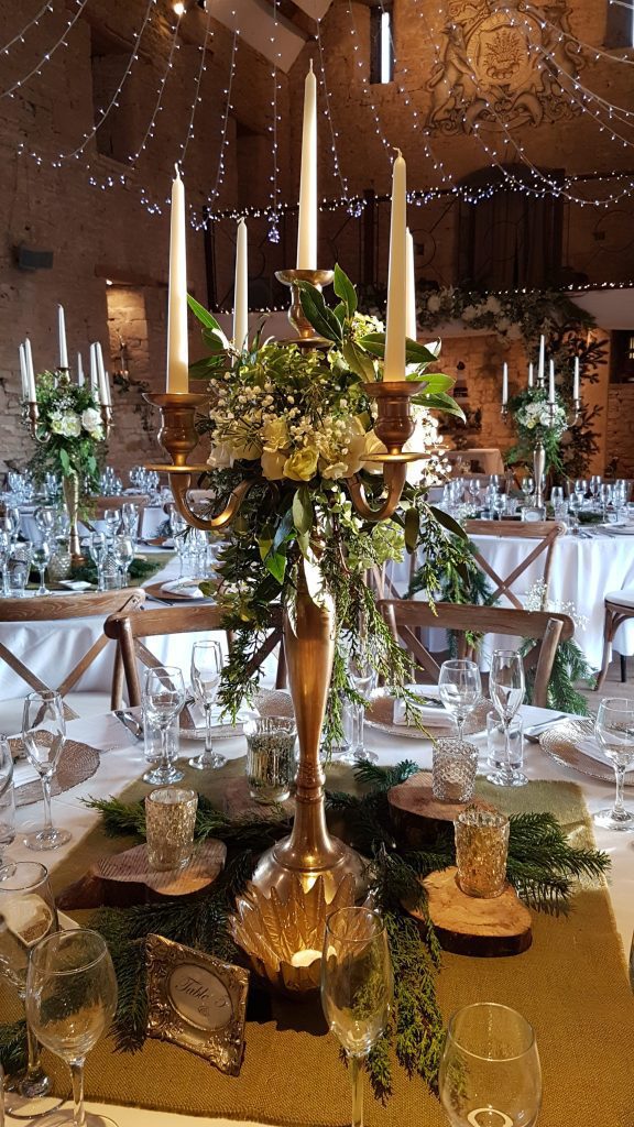 Gold candelabra, gold charger plates, and wooden slice with hessian table cloth and tealight holders in front of a white and green arch full of ivy and winter Christmas tree branches for a woodland winter wedding available to hire