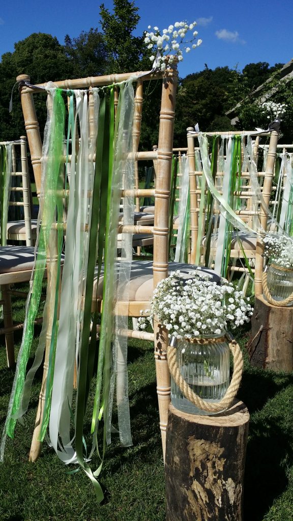 At Hyde barn in the Cotswolds Chivari chairs are set up for the wedding ceremony in the garden in front of the pergola each chair has a string of white and different shades of green ribbon tied across the back and a sprig of gypsophila on the aisle side. Also running down the aisle are tree stumps with a glass jar with a rope handle and a large bunch of gypsophila in it.