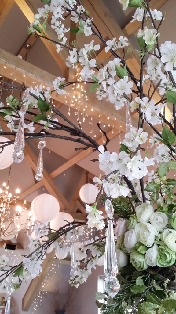 close up of Luxury quality faux flowers in whites and greens, with spring blossom branches of white blossom and clear crystal hanging from them. with a backdrop of fairy lights wrapped around beams in the eaves at Hyde Barn in the Cotswolds for hire
