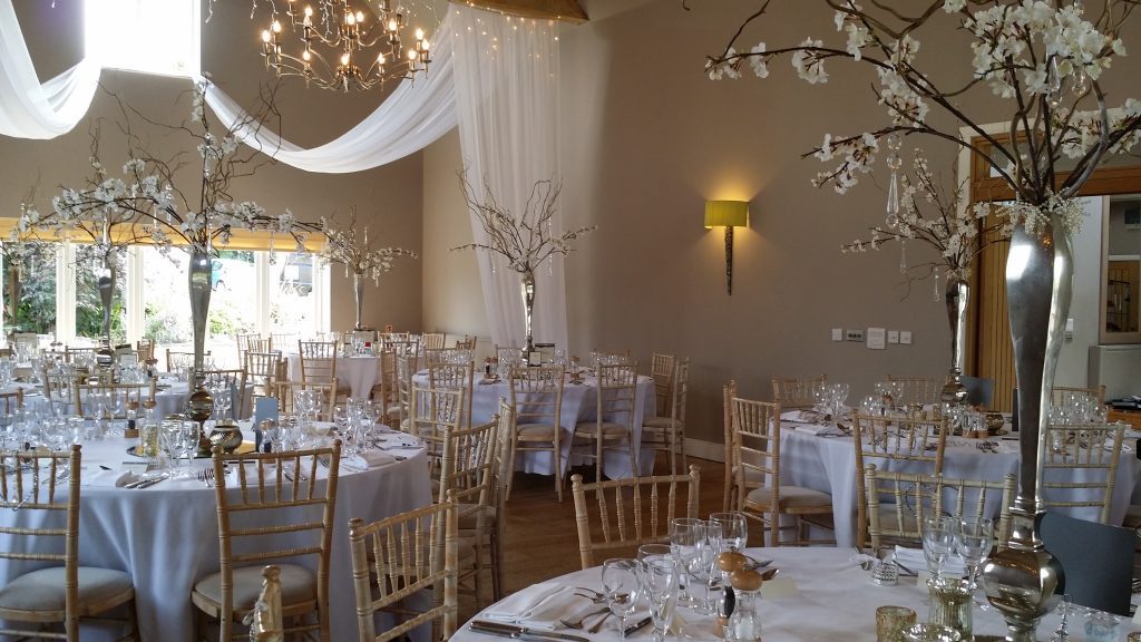 A room set up at Hyde Barn in the Cotswolds,  With many tables with white tablecloths set on round tables with a high shine silver vase on a round mirror with twisted willow and fake blossom branches coming from it, and hanging crystal scattered across the arrangement