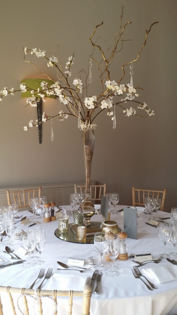 A table set up at Hyde Barn in the Cotswolds, on a white tablecloth set on a round table with a high shine silver vase on a round mirror with twisted willow and fake blossom branches coming from it, and hanging crystal scattered across the arrangement