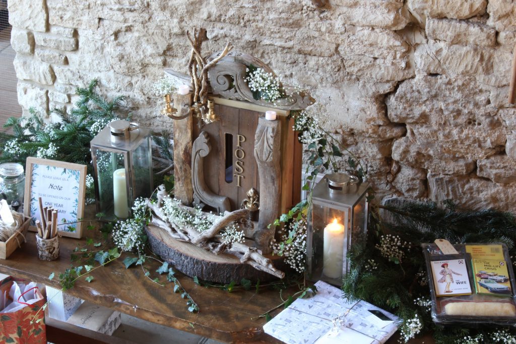 with a backdrop of Cotswold stone sits a Vintage rustic wooden crate post box with ivy and gypsophila, a silver lantern either side and winter pine branches, the guestbook and gift table available to hire at the Great Tythe Barn Tetbury