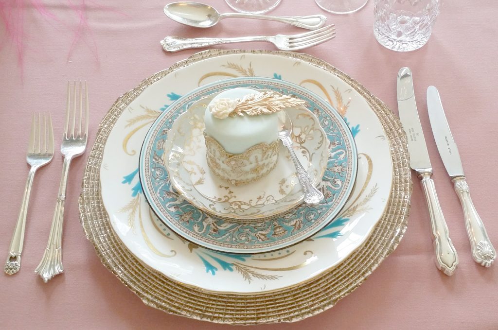 table place setting of a gold charger plate then starter main and dessert plate stacked on top in blues whites and gold with a mini iced cake say on the top and silver vintage mix and match cutlery for a starter , main course and dessert framing the plates on a pale pink tablecloth to hire
