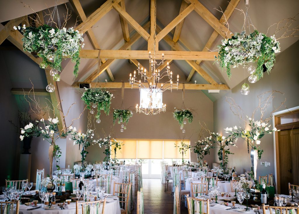 tall white and green wedding table centre decor with lisianthus ivy and twigs, surrounded by a hessian tablecloth and wooden slices as log tealight holders and green stemmed wine glasses at Hyde barn in the Cotswolds available for hire