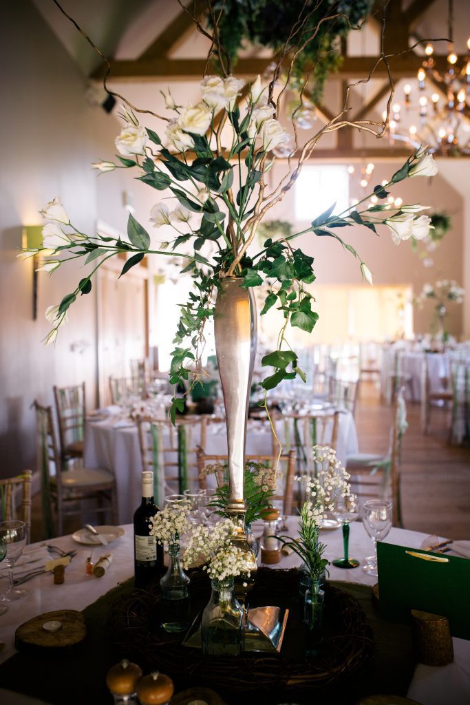 At Hyde barn in the Cotswolds a Large lisianthus and twisted willow silver vase centrepiece, with recycled glass bottles and gypsophila on a log round and log tealight holders to hire