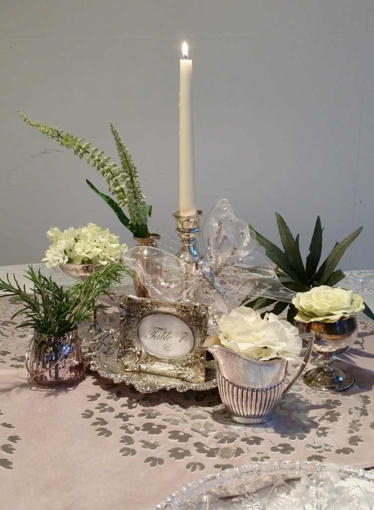 Simple centrepiece set on a vintage silver tray with a vintage Silver jugs and vases the middle and the table number in a silver ornate photo frame with poses of white roses and greenery and a silver candlestick and a lit white tapered candle