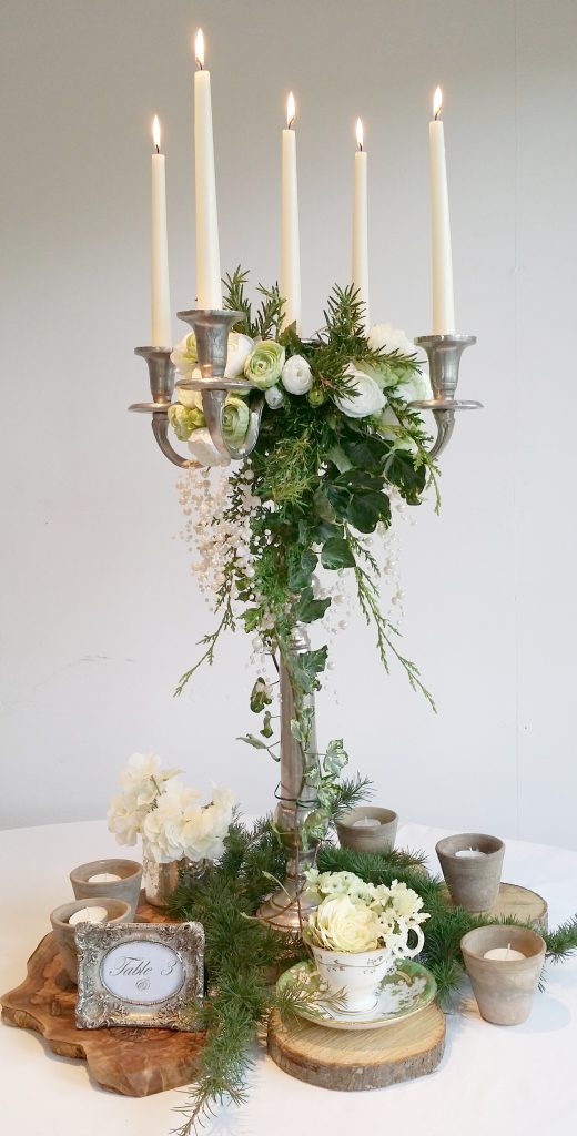 luxury silver candelabra with a garland of flowers at the centre and ivy and fake roses mixed in, around the base are tree trunk slices and tealight holders in between fresh pine available to hire for weddings and events