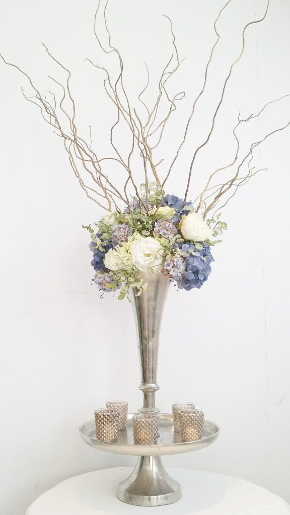 Large silver trumpet vase with tealights around the base and filled with blue and white hydrangeas and twisted willow available to hire