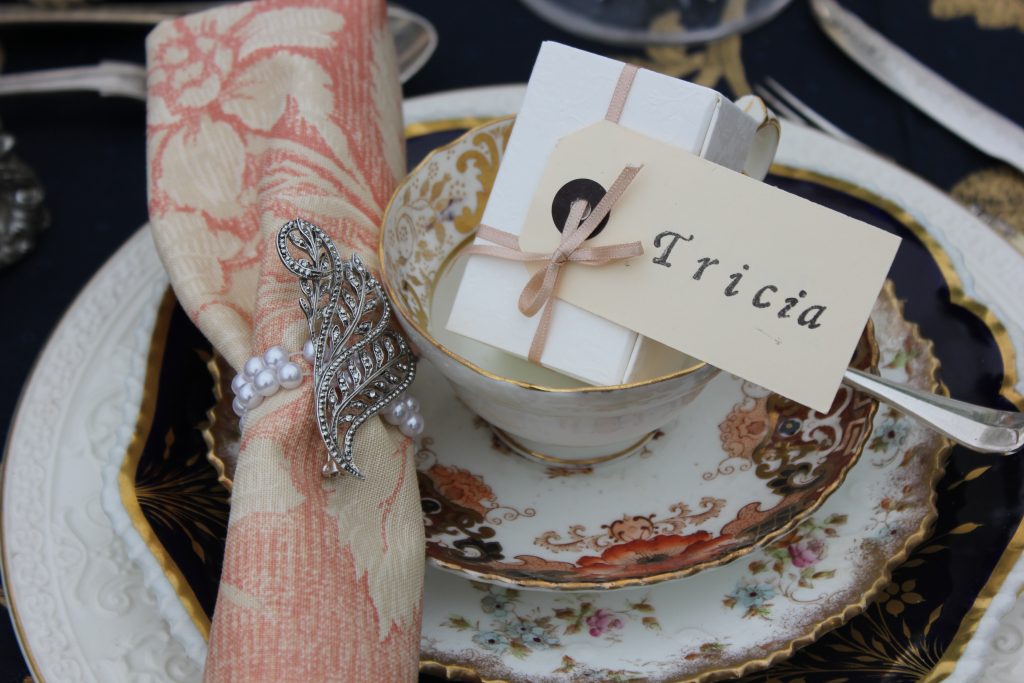 A close up of a vintage tea cup, saucer and plate with a box of favour n the cup and a pink napkins to the left with a silver diamante and pearl broach wrapped round it