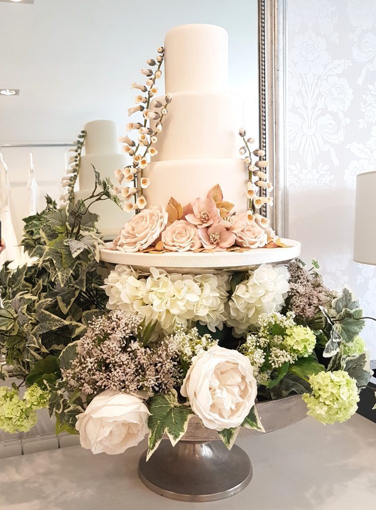a grand layered two tiered silver cake stand. The bottom layer is packed with fake white roses, ivy and hydrangeas the top layer is a three tier pale pink cake with a beautiful selection of edible roses around the bottom all available to hire