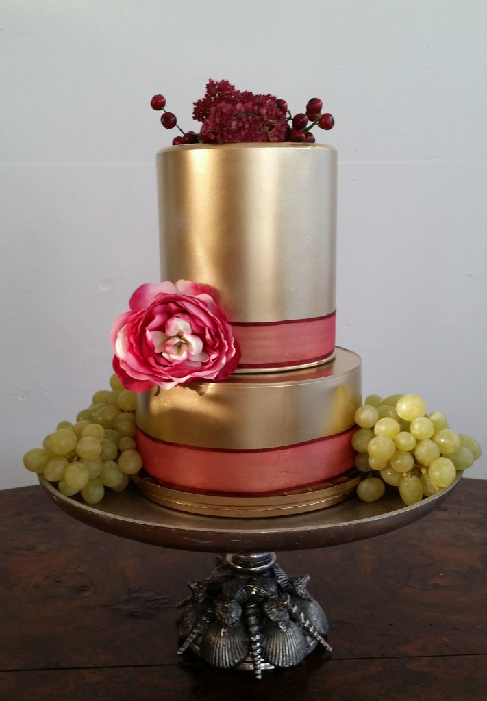 Sea shell silver pedestal cake stand with a two tier gold cake on top and a red ribbon wrapped round each layer to hire