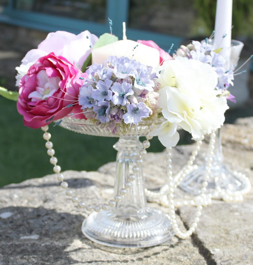a glass pedestal cake stand with a string or pearls hanging from it and bright pink and lilac faux flowers sat on top with a pillar candle in the middle