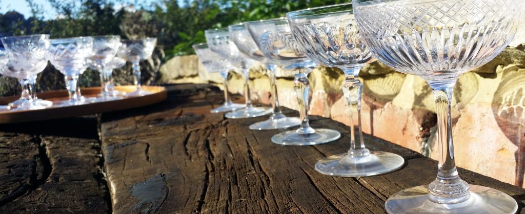 A line up of cut crystal vintage champagne saucers in front of a Cotswold stone wall reflecting the sun and blue sky, available to hire