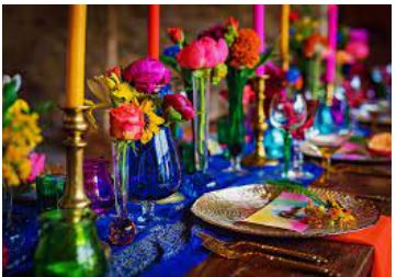 A beautifully bright and colourful table made up of  royal blue table runner on a trestle table, hosting brass candlesticks with multicoloured candles and a variety of coloured glass vases filled with many colourful flowers and place settings of gold charger plates and laid up with silver vintage cutlery
