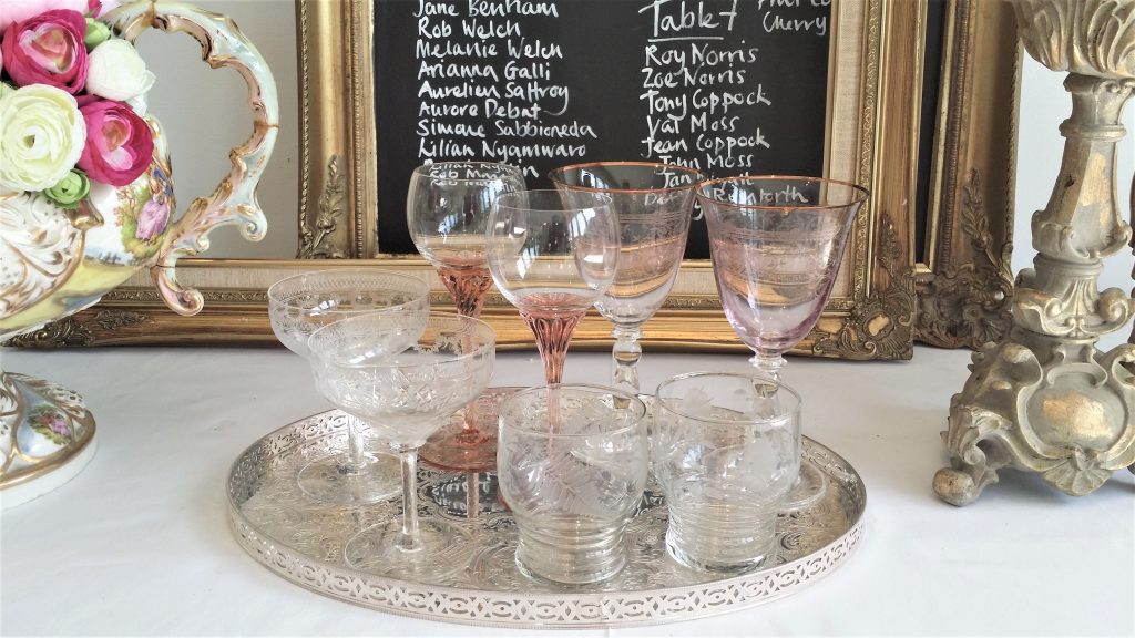 on a silver vintage tray there are vintages pink coloured wine glasses, embossed champagnes saucers and cut glass water tumblers in front of a gold table plan for hire