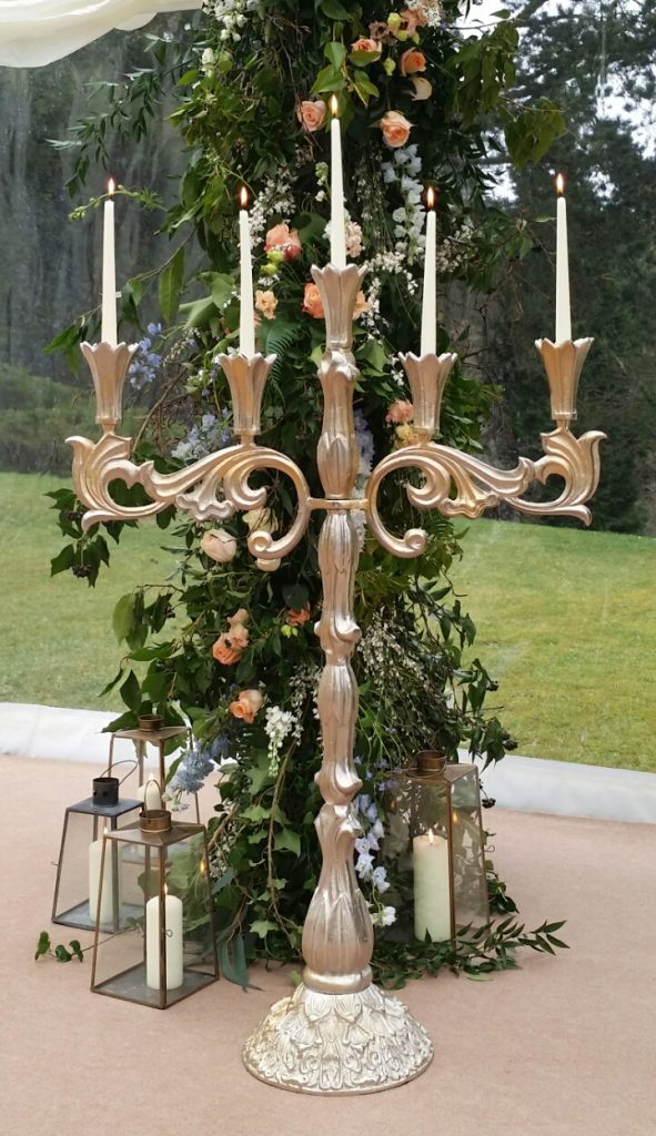 Stunning ornate extra large candelabra with pillar candles in front if a large floral arch and glass lanterns around the base for hire