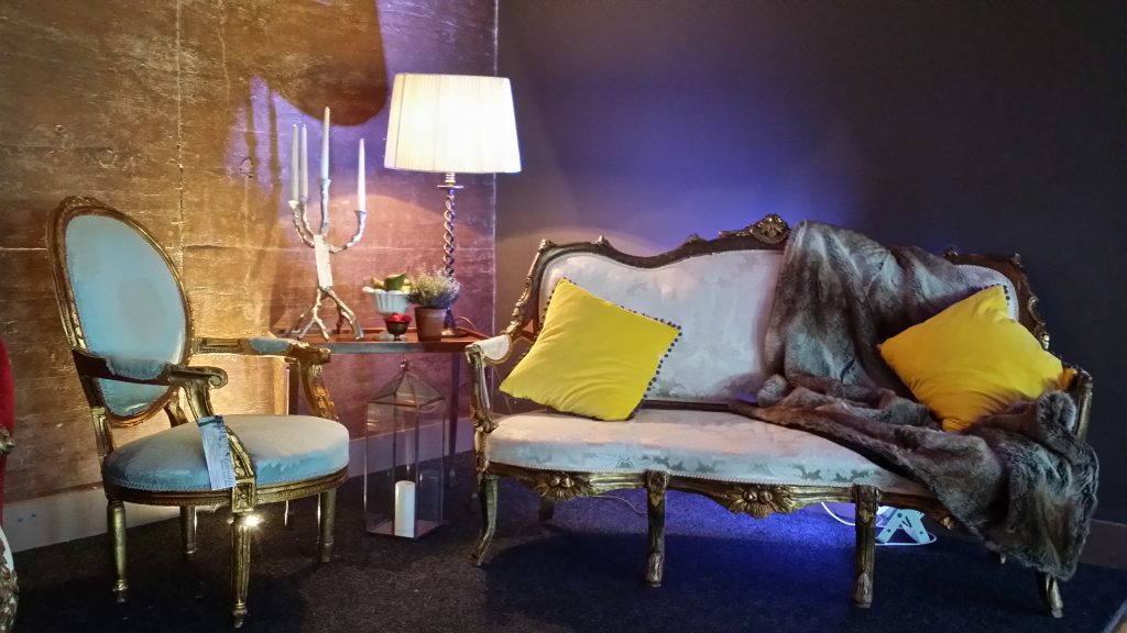 White and gold rococo vintage sofa with a fur through and two mustard scatter cushions with a light blue chair with a gold frame to the left and a twig candelabra and lampshade behind with a blue lighting, all available to hire