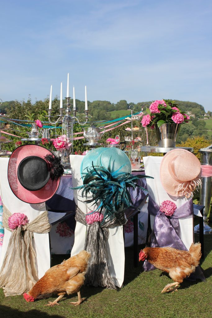 a table set up for a garden party of afternoon tea with vintage crockery and tableware and candelabras and 3 tiered cupcake stands. on the back of three chairs are funky hats, one bright pink, one blue with feathers and one baby pink