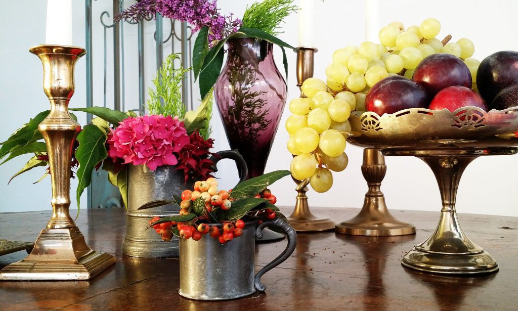 close up of three brass candlesticks and a brass vintage pedestal cake stand and two pouter mugs and in the back a purple vase, all filled with an array of grapes and plums, berries and flowers in reds and pinks to contrast amongst greenery and leaves