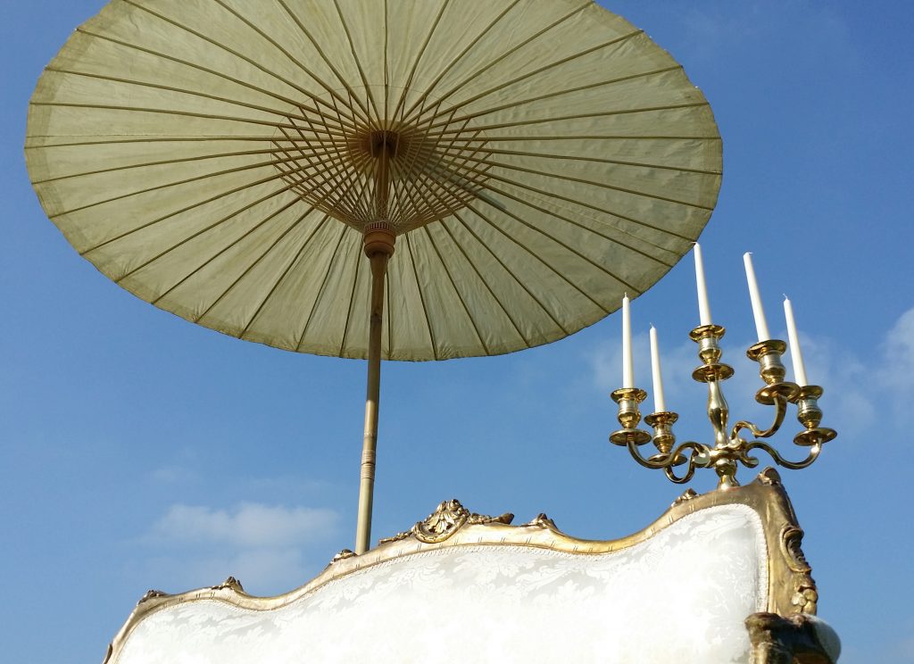 A white vintage sofa with an ornate gold frame surround, arms and legs set up on the grass with a large candelabra and Japanese style paper parasol with bamboo spokes behind it