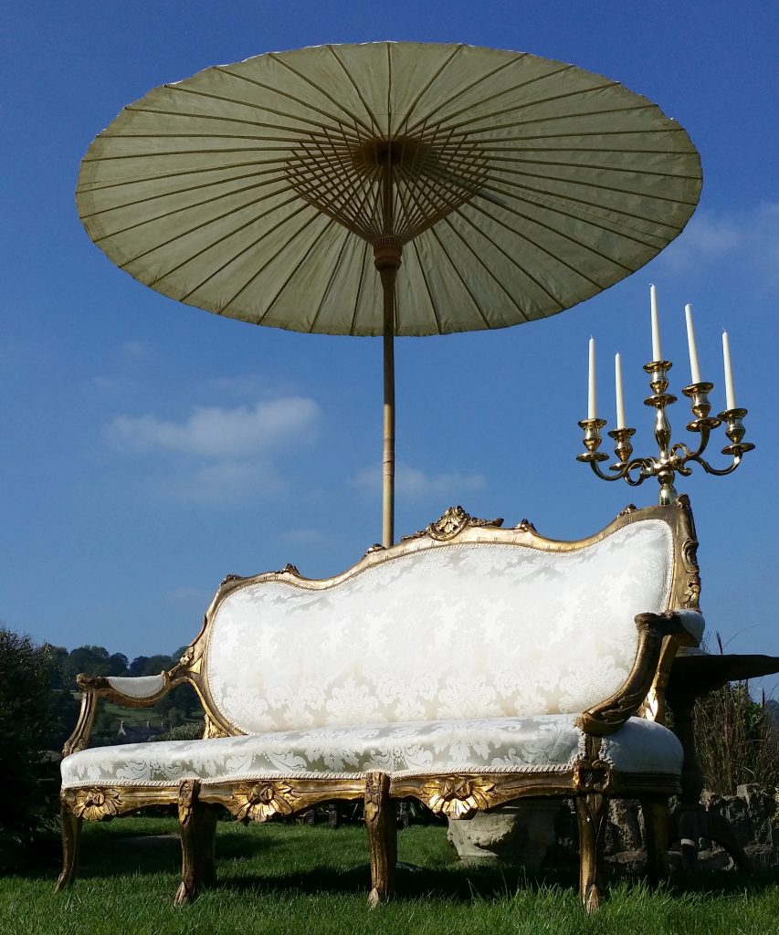 A white vintage sofa with an ornate gold frame surround, arms and legs set up on the grass with a large candelabra and Japanese style paper parasol with bamboo spokes behind it