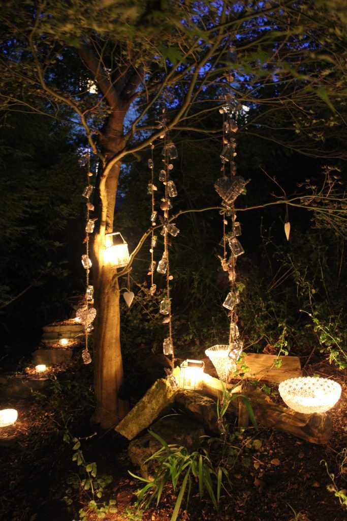 outside in the woods set on stumps are cut crystal vases with tealights in them creating a warm glow with mini jars on a string hanging from the trees reflecting the candle light to hire