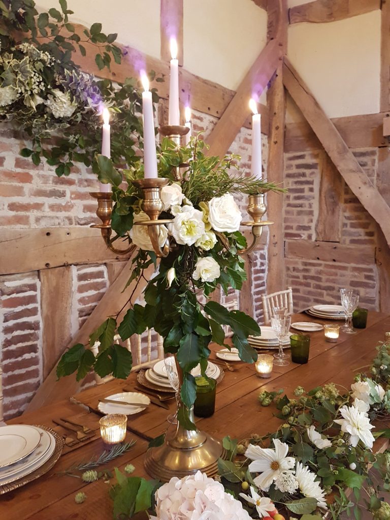 Stone barn with trestle tables and white flowers and greenery wrapping a gold candelabra with a white and green garland and table runner in the front for hire