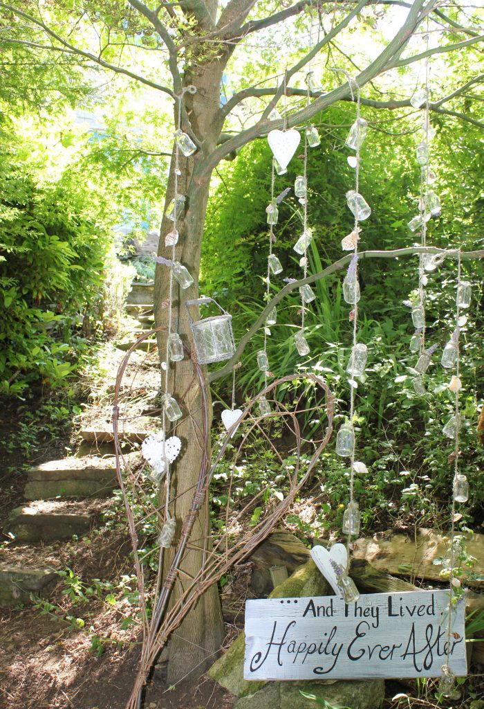 from a tree branch is hanging string of mini jars with shells and knots for stem flowers and a, And they lived happily ever after sign underneath, with a wicker heart leant up against the tree trunk available for wedding and event and prop hire