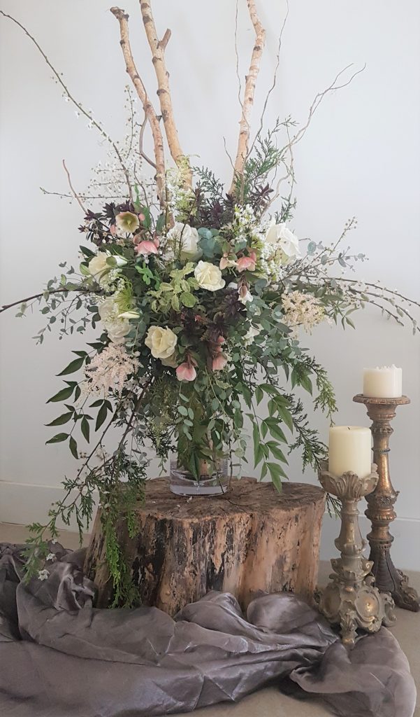 floral arrangement in a vintage vase sat on a tree stump with two gold Blenheim ornate candlesticks with pillar candles next to it
