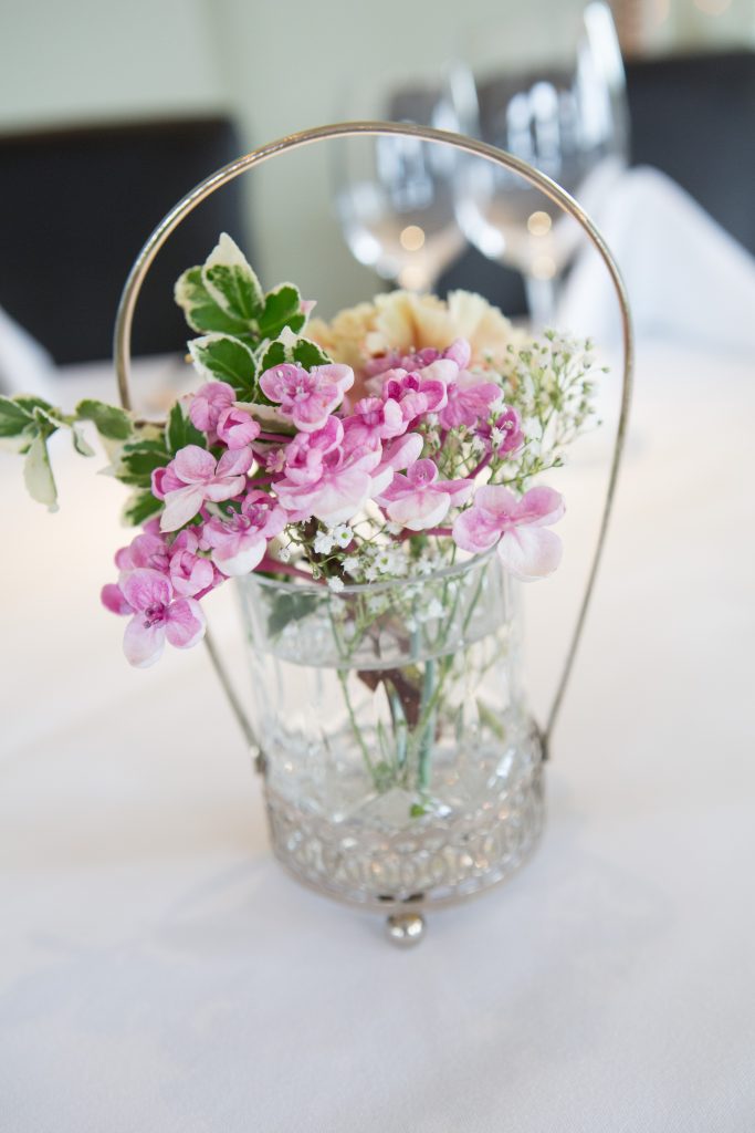 A vintage cut crystal glass bud vases filled with antique pink roses, pink hydrangea,  white daisies and gypsophila and sprigs of fresh eucalyptus and a silver hanging heart with crystals
