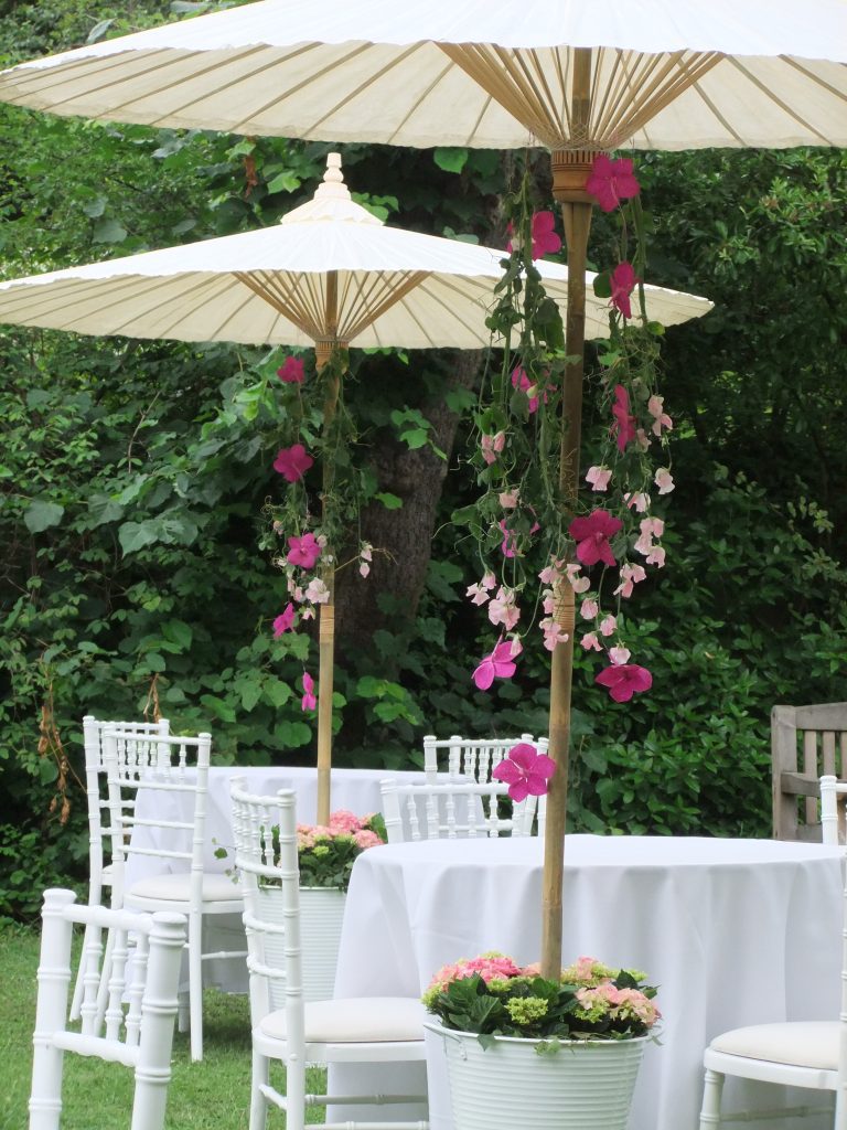bamboo parasols for summer wedding hire with flowers Celebration of life garden set up, Next to the table stands a white bucket filled with pink hydrangeas and greenery covering the base of a beautiful Japanese style paper and bamboo parasol with strings of Hot pink orchids and baby pink sweet peas hanging from the spokes