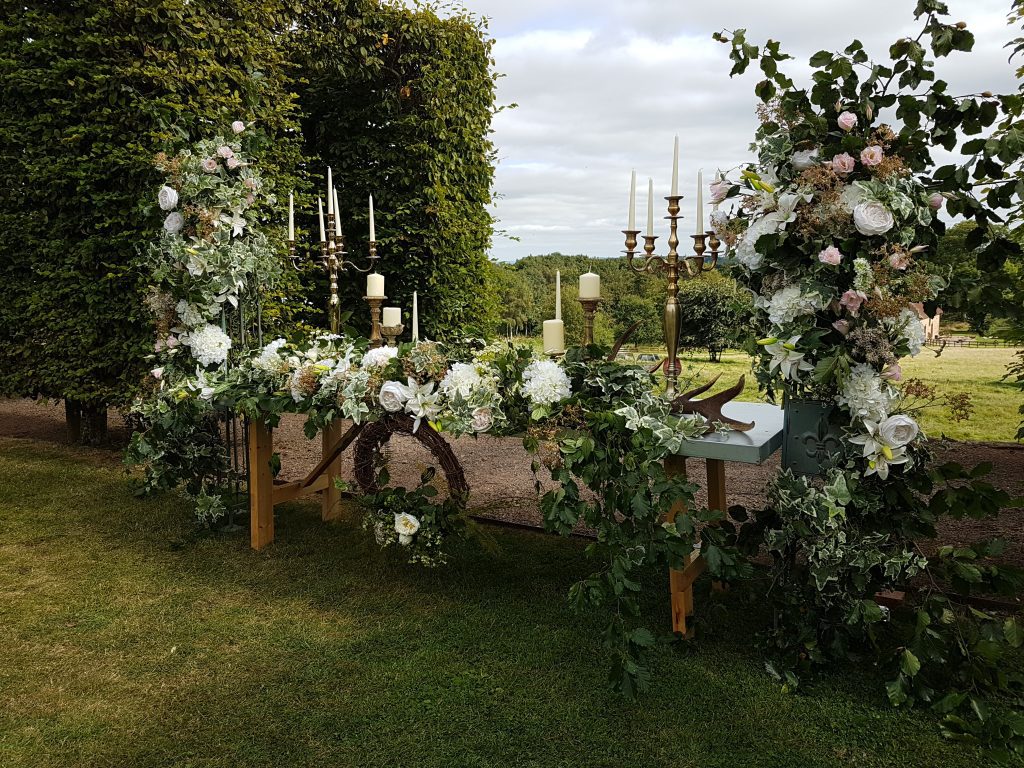 A trestle table with a garland of faux and fresh white flowers and greenery, with gold candelabras and a wicker wreath hanging in front, either side are two pillars covered with quality fake flowers such as roses, hydrangeas and lilies and greenery.