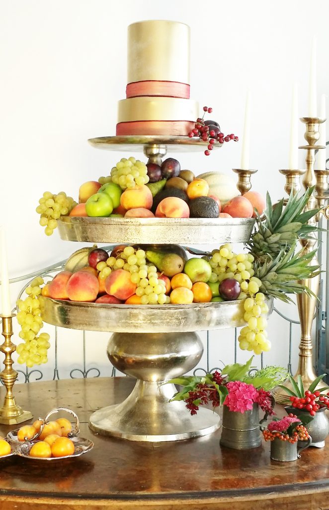 a grand layered three tiered silver cake stand. The bottom and middle layers are filled with grapes, peaches, apples and pears the top layer is a gold frosted two tier cake with a red ribbon around the bottom of each layer all available to hire