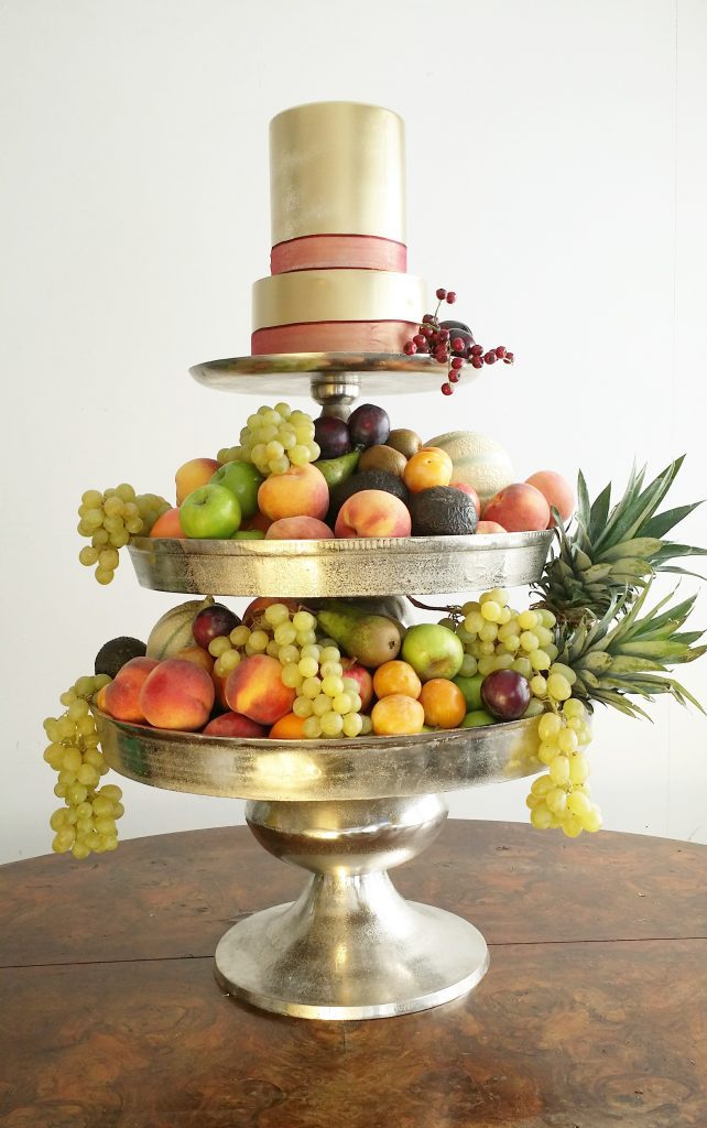 a grand layered three tiered silver cake stand. The bottom and middle layers are filled with grapes, peaches, apples and pears the top layer is a gold frosted two tier cake with a red ribbon around the bottom of each layer all available to hire