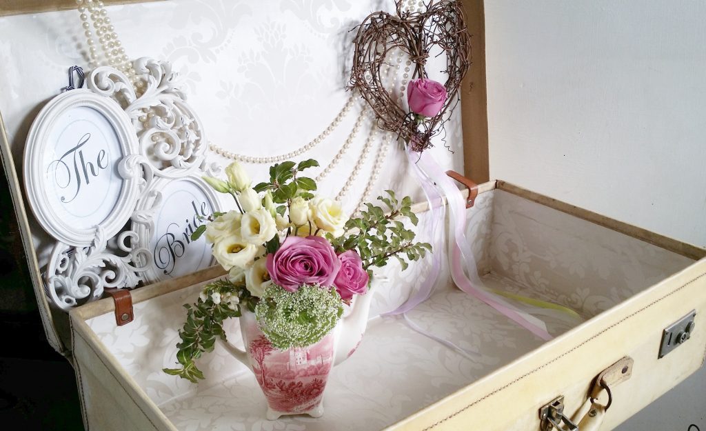 a vintage beige or cream suitcase for cards or wedding gifts with a vintage teapot filled with fresh flowers and pearls hanging across the bank and a wicker hanging heart hanging from the top with a single pink rose at the bottom for hire
