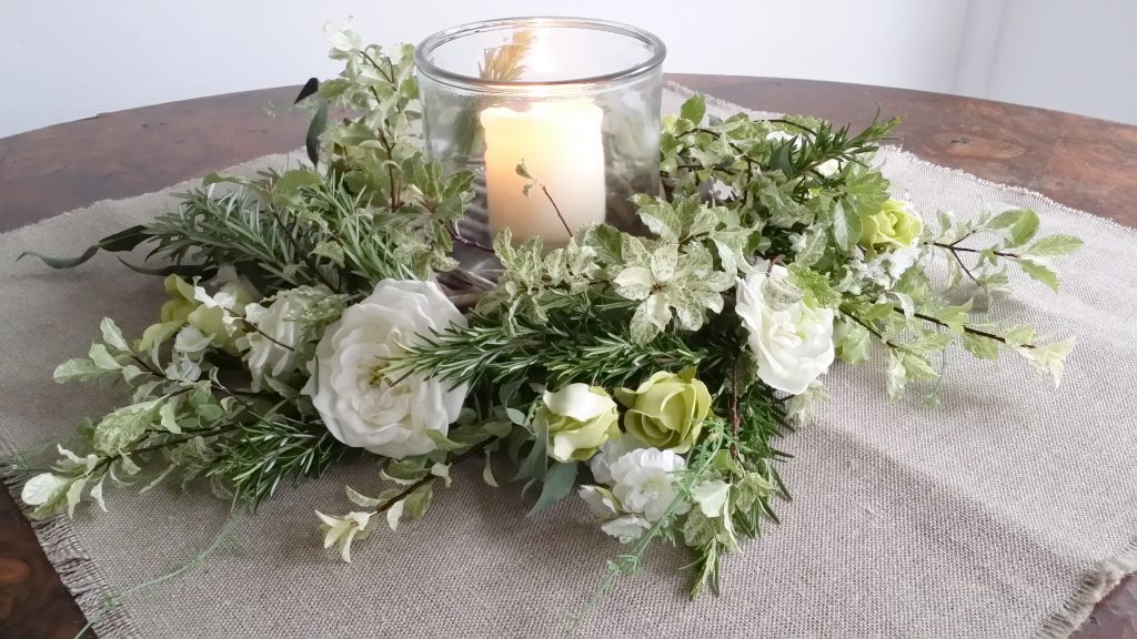 simple centrepiece of a square of hessian tablecloth with a fake floral wreath and fresh greenery mixed in sat around a glass vase with a church candle in it for hire