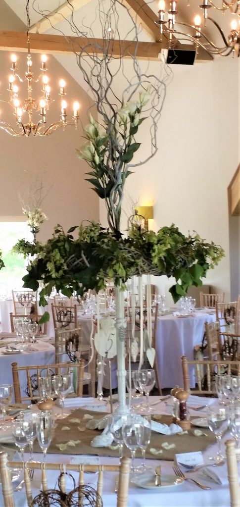 At Hyde barn in the Cotswolds, A table centrepiece of a white central pillar with a large wreath of fresh greenery at the top, with twisted willow and white lisianthus coming from the centre reaching to about five feet high. this arrangement is sat on a square hessian cloth in the centre of a round table covered in a white tablecloth.