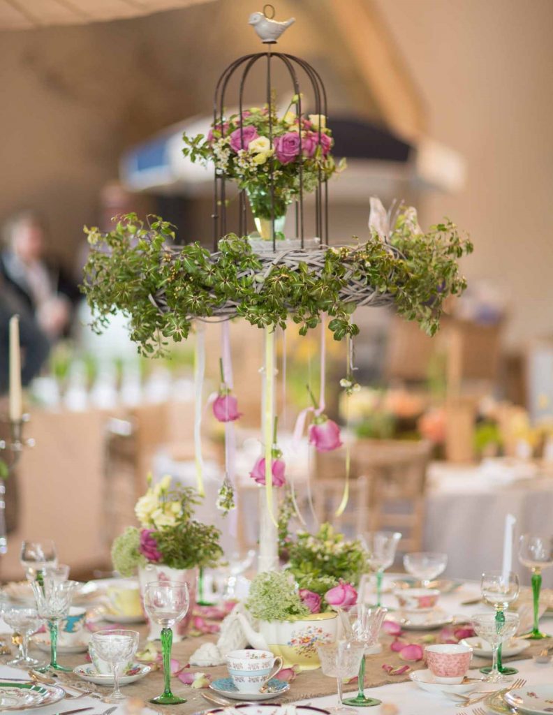 garden party afternoon tea with vintage tea cups and saucers with a centrepiece of a large white stand with a wicker wreath on top filled with greenery and a bird cage on top with a vintage bud vase filled with roses for hire as a centrepiece