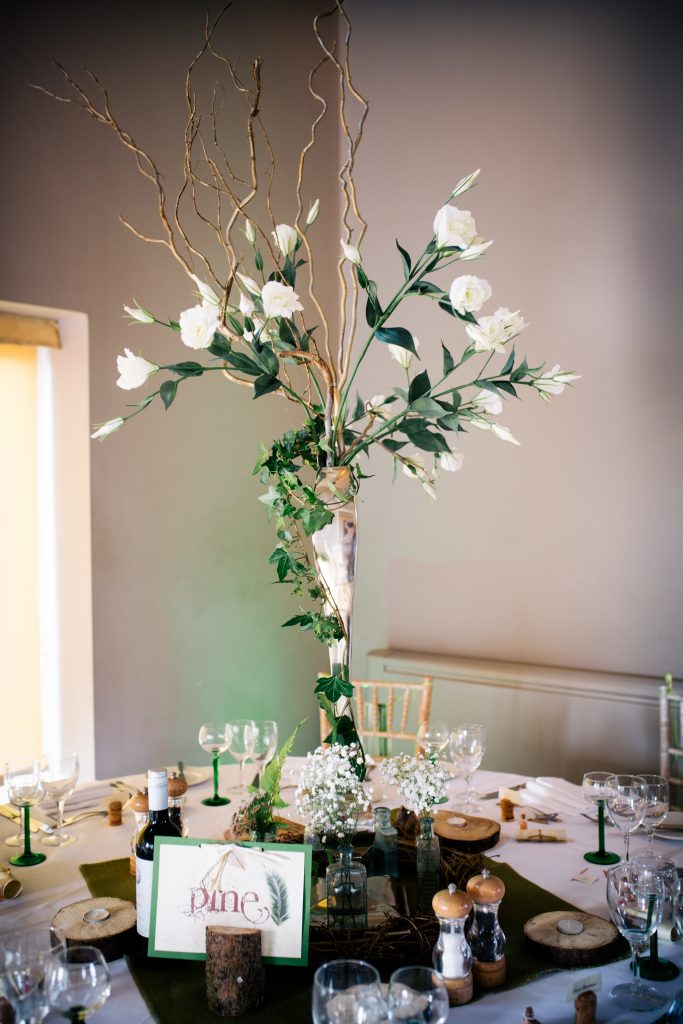 tall white and green wedding table centre decor with lisianthus ivy and twigs, surrounded by a hessian tablecloth and wooden slices as log tealight holders and green stemmed wine glasses at Hyde barn in the Cotswolds available for hire