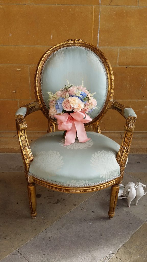 A vintage light blue embroidered chair with a gold frame and a bouquet of blue, pink and white roses placed on it and a pair of white bridal shoes on the floor to the right furniture available to hire