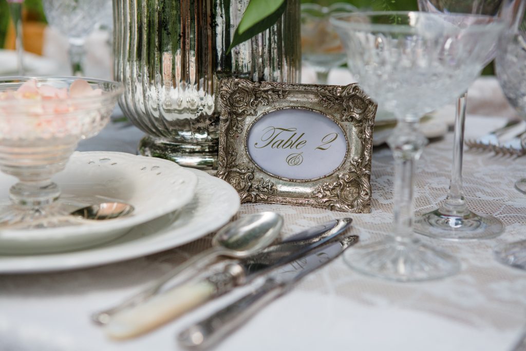 Close up Of a silver ornate photo frame with table two in it for a wedding table plan and vintage silver cutlery and glass sundae bowl with cut crystal champagne saucer to the right hand side to hire