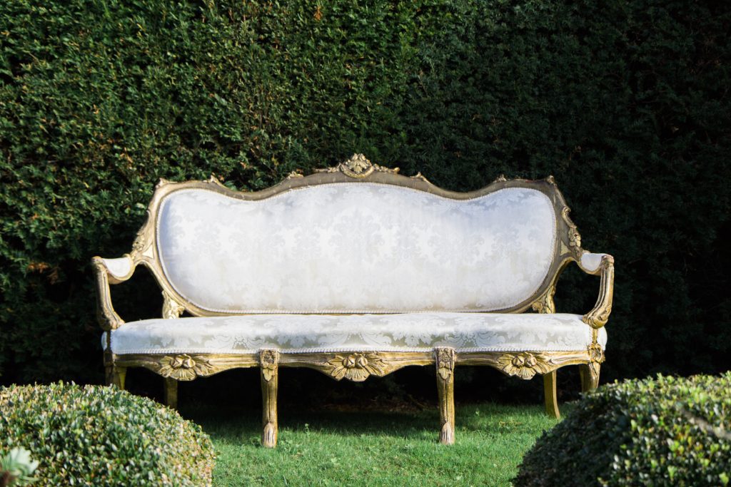 Stunning vintage rococo white and gold ornate sofa in front of a pruned hedge at Sudeley castle for hire