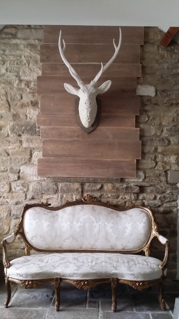 White and gold ornate sofa beneath a large white stag head prop and backed on wooden boards on a Cotswold stone wall, with much more vintage furniture available in the Cotswold's