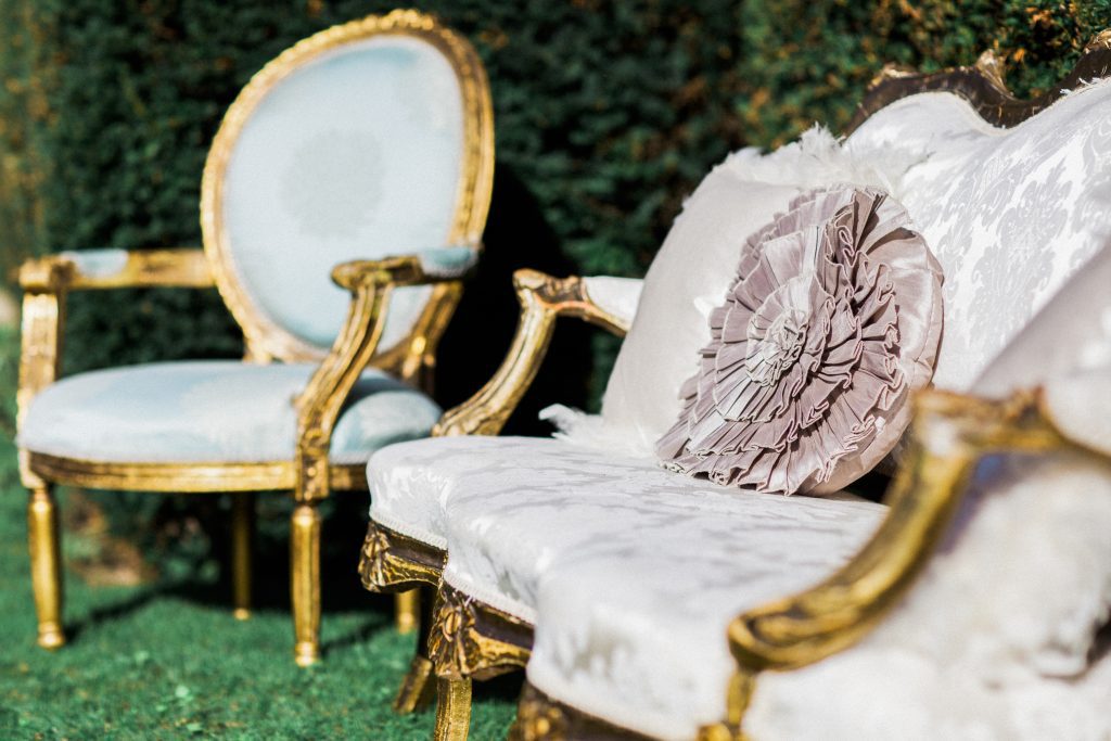 A white vintage sofa with an ornate gold frame surround, arms and legs and a blue upholstered chair with a gold frame in front of a hedge