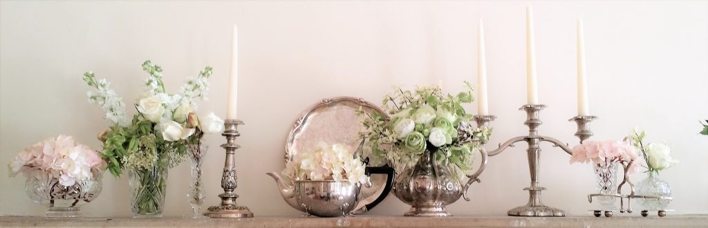 vintage glass vases and silver teapots with fake flowers in sat on a mantlepiece with a silver candlesticks and a silver candelabra mixed between the arrangements for hire