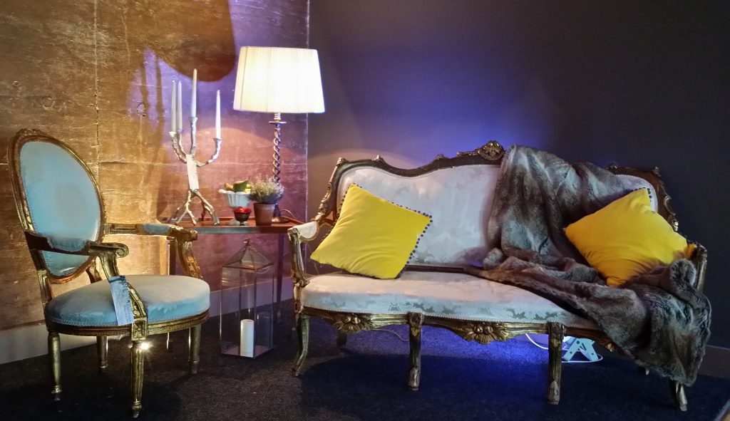 White and gold rococo vintage sofa with a fur through and two mustard scatter cushions with a light blue chair with a gold frame to the left and a twig candelabra and lampshade behind with a blue lighting, all available to hire