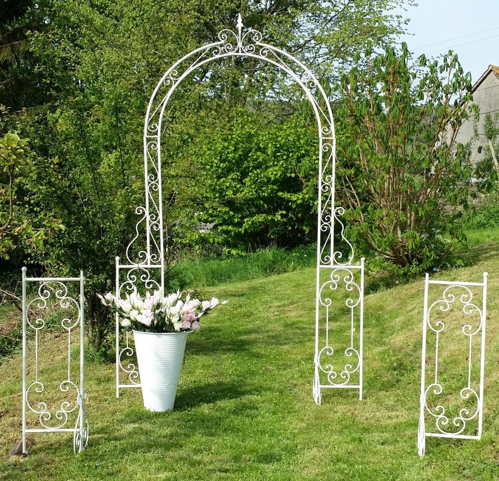 extra large white wedding arch with twisted details set up for and aisle outside with a white bucket of faux flowers for hire